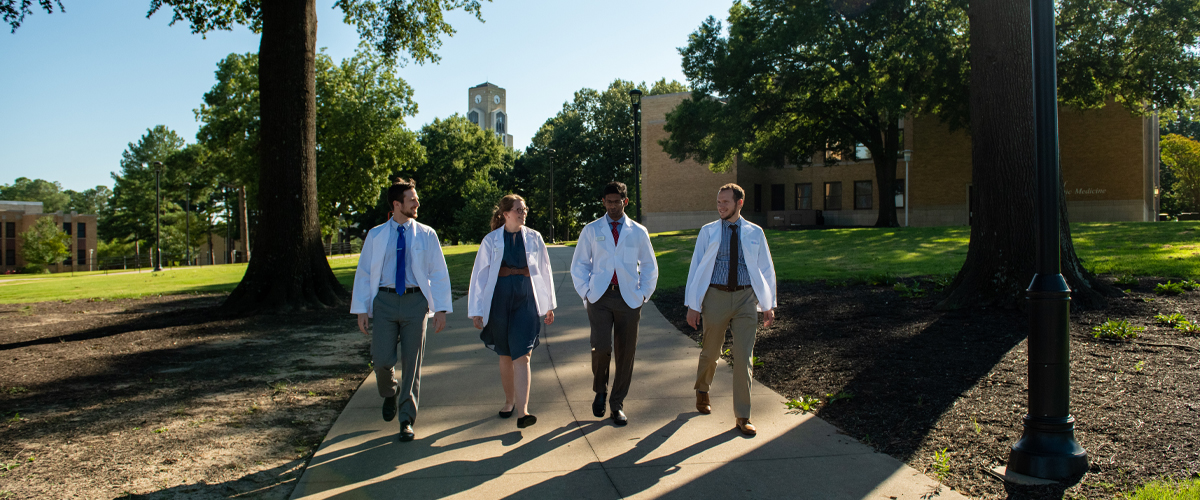 A group of 4 NYITCOM medical students with white coats walking on the Jonesboro, AR campus.