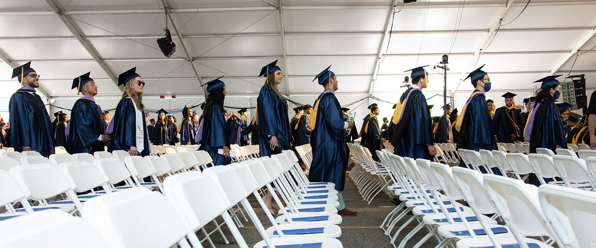 Procession of students inside large tent at Commencement ceremony. Foreground is full of chairs.