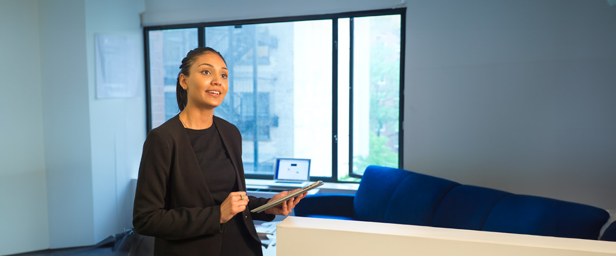 A woman wearing business attire in a classroom holding a tablet.