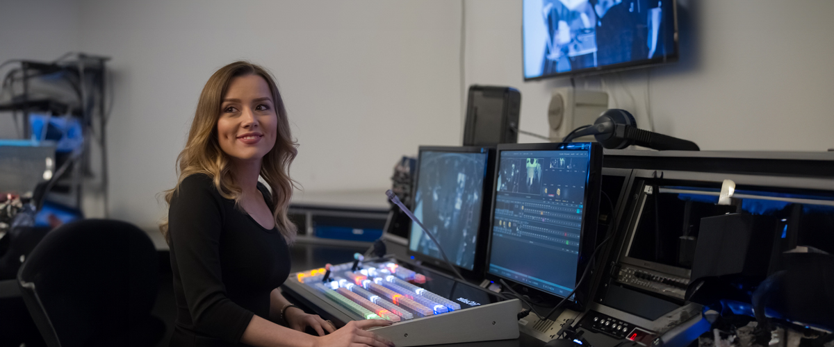 A student works in a production studio with two screens and a switch board.