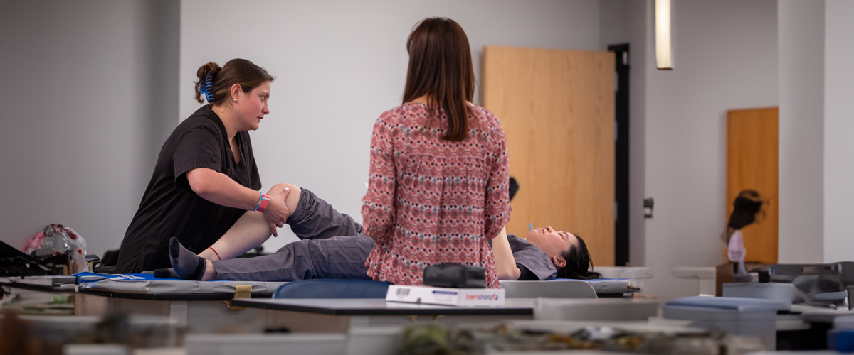 A physical therapy student works on another student's leg mobility on a table.