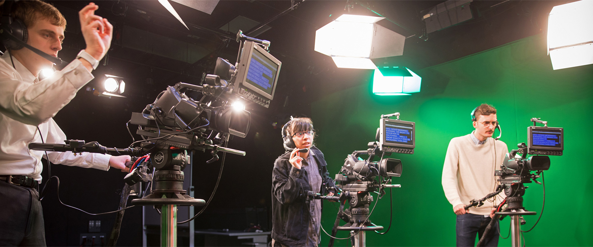 A student works in a television studio with cameras and a green screen.