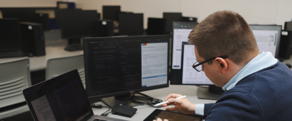 Man at a desk with laptop and computer screens looking at an iPad