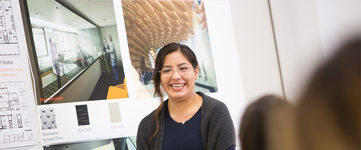 An interior design student presents in front of large posters in a classroom.