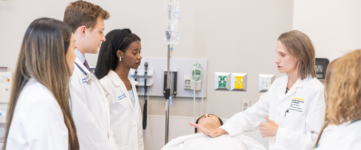 Three medical students in white coats observing a demonstration in a simulation lab led by an instructor.